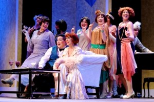 Lea DeLaria and Andrea McArdle seated on sofa surrounded by other cast members dressed for Roaring 20’s partying. (Photo credit: Mandee Kuenzle)